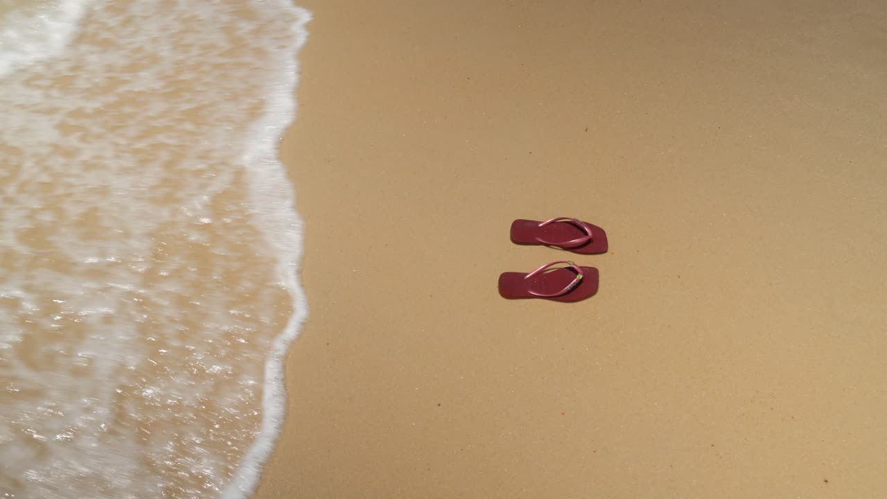 Primo piano di sandali infradito Havaianas rossi adagiati sulla sabbia bianca mentre la marea in arrivo li trascina fuori dall'inquadratura. Ripreso sulla spiaggia di Ipanema, Brasile