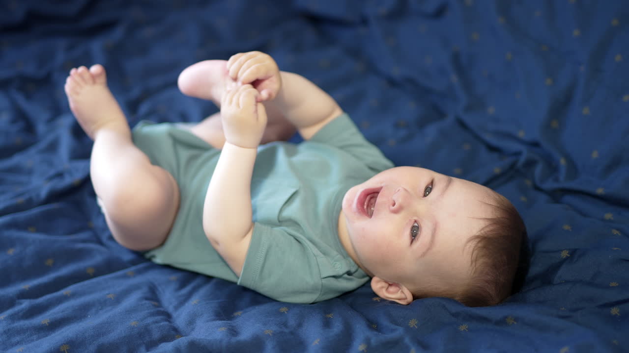 Healthy beautiful kid playing with his feet holding them up. Adorable child laughing cheerfully. Close up. Blue backdrop.