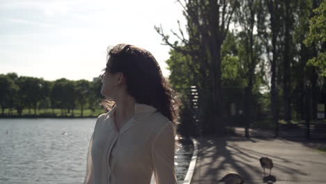 portrait of a fashionable and beautiful italian model walking in a tranquil park near a lake, wearing a transparent shirt, looking around and having fun in london, united kingdom