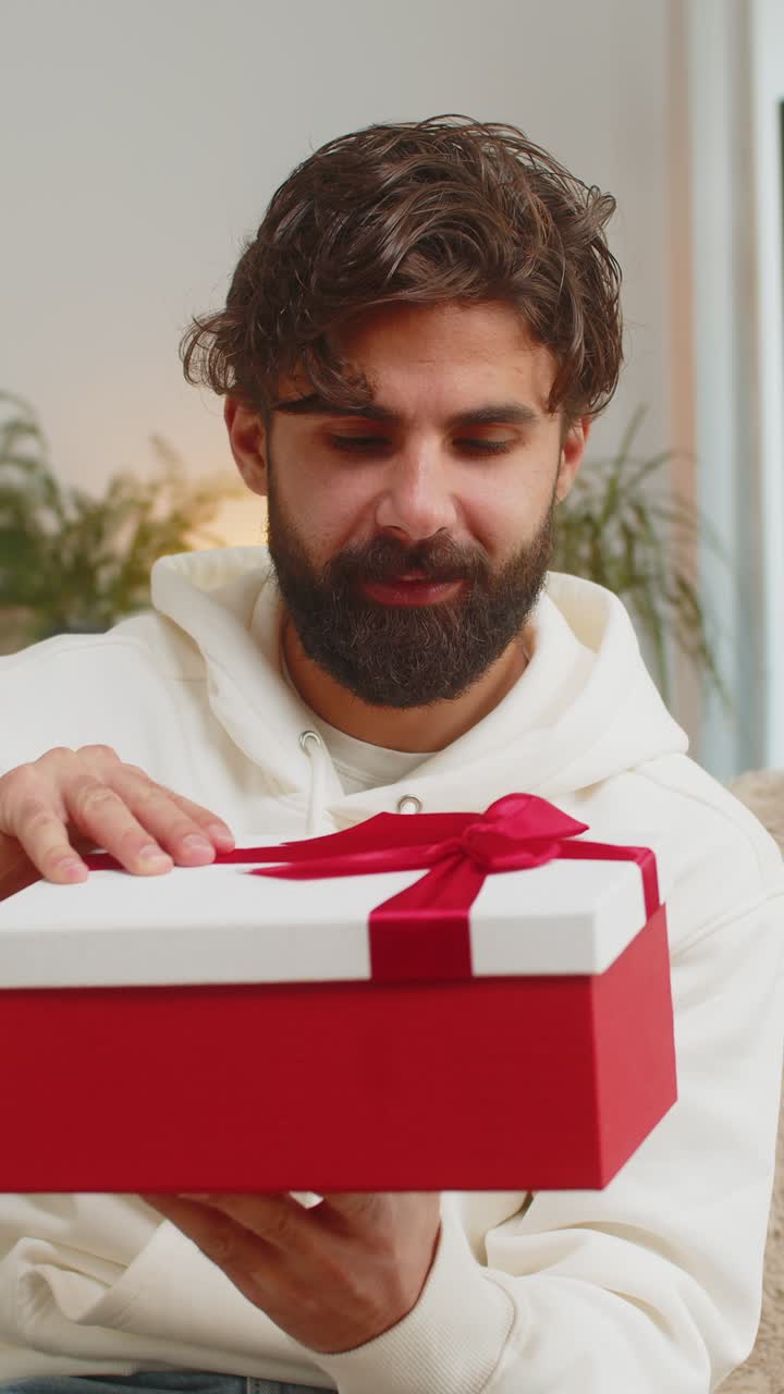 Un giovane indiano felice che tiene in mano una scatola di regali di compleanno con un nastro che lo congratula seduto sul divano a casa