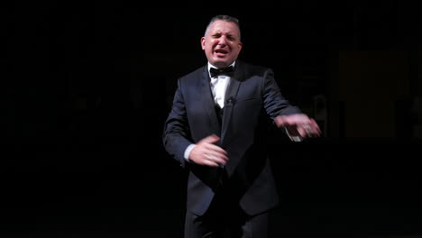 man is speaking in the light of the floodlights standing on stage to the camera. actor, artist on performance. activ gestures with hands.