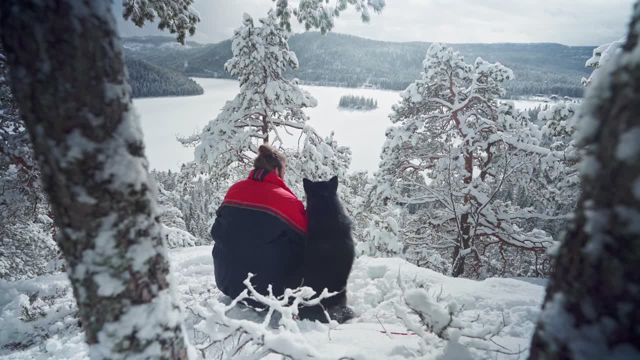 Vista trasera de un hombre con su perro Alaskan Malamute sentado en una montaña nevada en Noruega - plano general.
