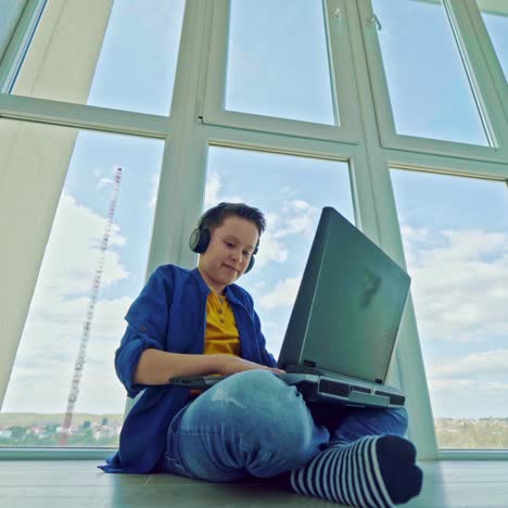 happy boy wins a game on a laptop. joyful teenager in headphones sitting by the window and playing video games on his laptop. gaming concept.