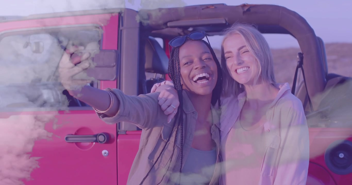 Due amiche che sollevano lo smartphone e inquadrano un selfie accanto a una Jeep rossa, immortalando un ricordo di un viaggio on the road