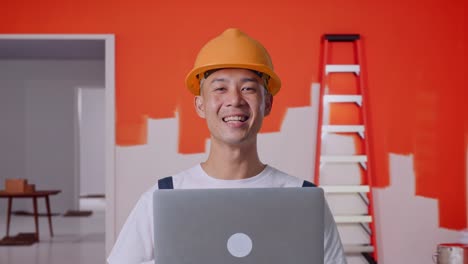 Close Up Of Asian Man Painter Wearing Safety Helmet Using A Laptop And Smiling While Standing With Painting Wall In Room