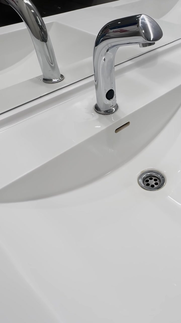 Close-up of a chrome automatic faucet over a white bathroom sink, with modern basin, drain, overflow slot, and clean public restroom design.