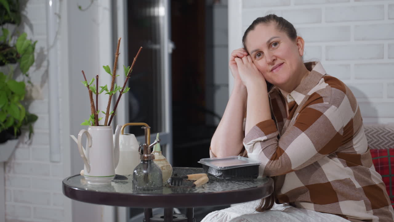 Portrait of woman resting head on hands at glass table surrounded by potted plants against brick wall backdrop in cozy home setting, smiling warmly and enjoying relaxing indoor gardening vibe