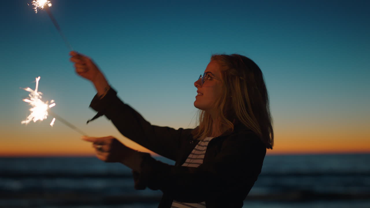 섣달 그믐날을 축하하며 해변에서 폭죽과 함께 춤추는 여자 바다에서 독립 기념일 축하를 즐기는 폭죽 불꽃놀이와 함께 즐겁게 춤을 추는 소녀