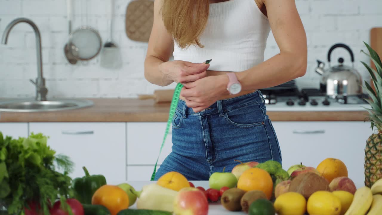 La ragazza si misura la vita in cucina vicino al tavolo con verdura e frutta.