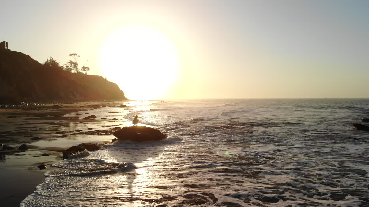Um homem em silhueta ao nascer do sol observando as ondas do oceano quebrarem na praia, com um drone recuando para revelar a deslumbrante costa de Santa Bárbara, Califórnia.