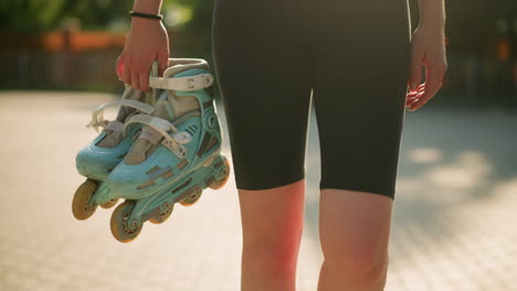 close-up of lady wearing black short walking outdoors holding cyan roller skates in her right hand under bright sunlight, blurred background features colorful greenery