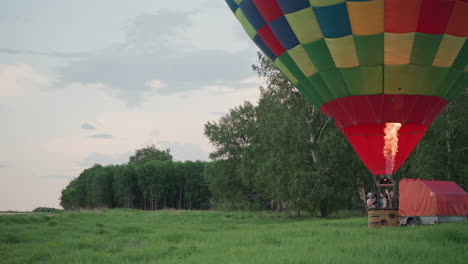 people riding in basket of hot air balloon while burner flame heats checker canopy over lush green field near forest at sunset with passengers watching sky as balloon slowly ascends
