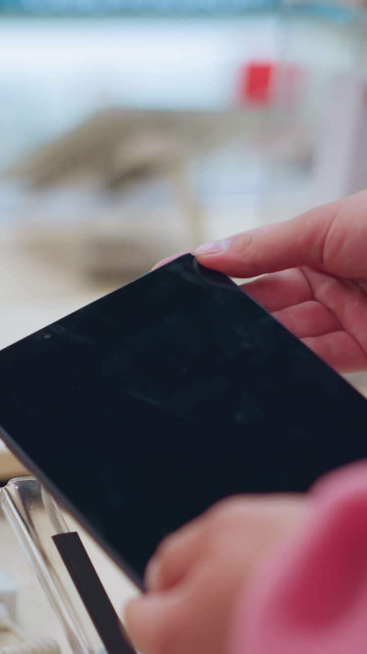 여자가 전자제품 가게에서 태블릿을 들고, 태블릿은 전기 케이블로 연결되어 있으며, 다른 태블릿들은 테이블 위에 끔하게 배열되어 있으며, 그녀가 화면을 관찰할 때 부드럽게 들고 있습니다.