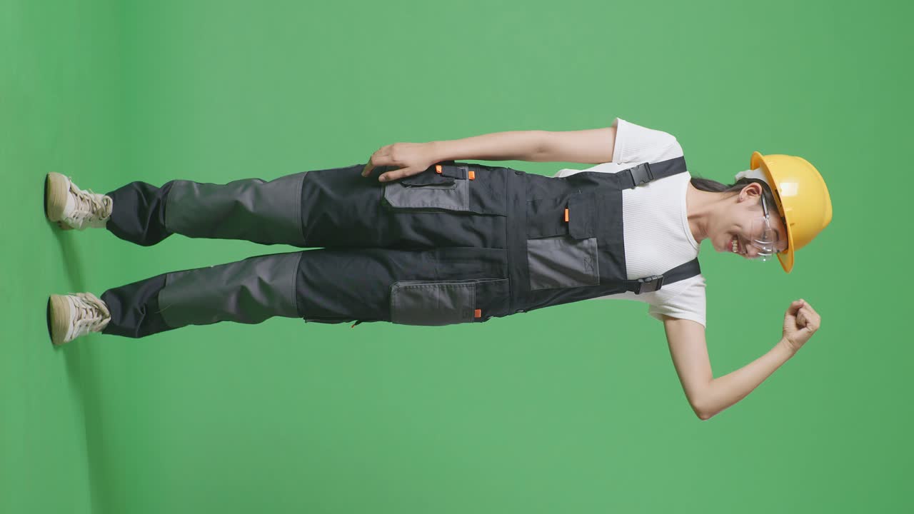 안경과 안전 헬을 입은 아시아 여성 노동자의 몸이 가득 차 있으며, 녹색 스크린 배경 스튜디오에 서 있는 동안 미소 짓고 팔꿈치를 구부리고 있습니다.