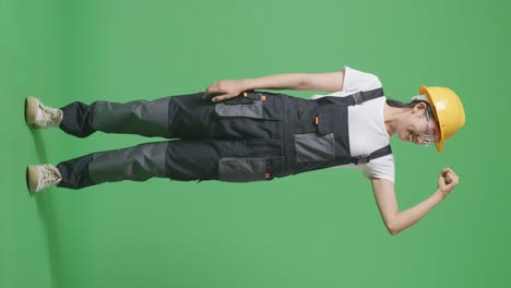 cuerpo lleno de mujer asiática trabajadora con gafas de protección y casco de seguridad sonriendo y flexionando su bíceps mientras está de pie en el estudio de fondo de la pantalla verde