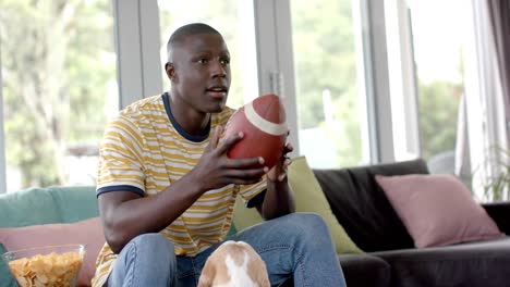 Happy-african-american-man-on-sofa-watching-rugby-match-at-home,-with-his-pet-dog,-slow-motion