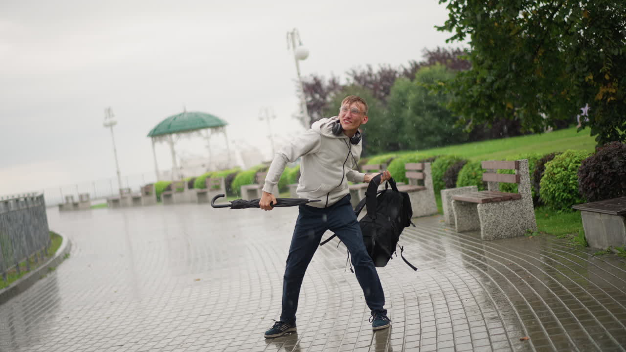 흰색 옷을 입은 남자가 백팩과 우산을 들고, 거센 바람과 싸운 후 장비를 되찾고 기뻐하며 점프하고, 즐거운 미소를 짓고, 물가의 벤치와 가로등이 보이고, 흐린 하늘을 반영하는 젖은 조약돌 길이 보이는, 솔직한 안도감