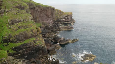 looking up from a rocky bay and out into the vast atlantic ocean as waves gently lap against a tall sea cliff in the ocean while seabirds fly around the grassy clifftops