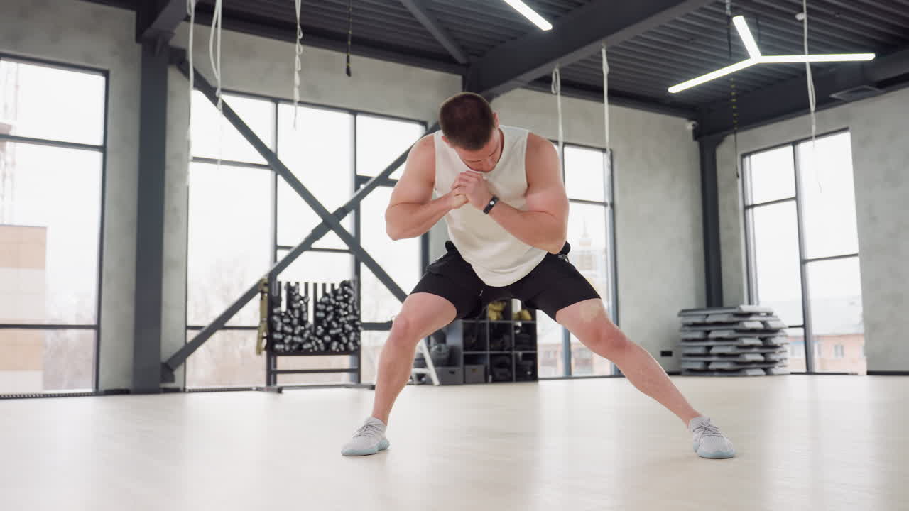Fitness freak performing side lunge warm up exercise in bright modern gym with white tank and black shorts balancing on light wooden floor beneath hanging straps and large windows urban