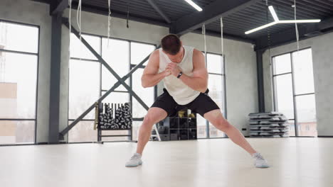 fitness freak performing side lunge warm up exercise in bright modern gym with white tank and black shorts balancing on light wooden floor beneath hanging straps and large windows urban