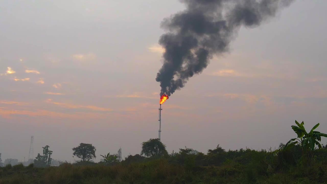 Fumo denso e fiamme vivaci si sprigionano dalla cima di una grande ciminiera industriale.