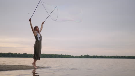 a young hippie woman in a dress and with feathers on her head makes huge soap bubbles at sunset on the shore of a lake in slow motion