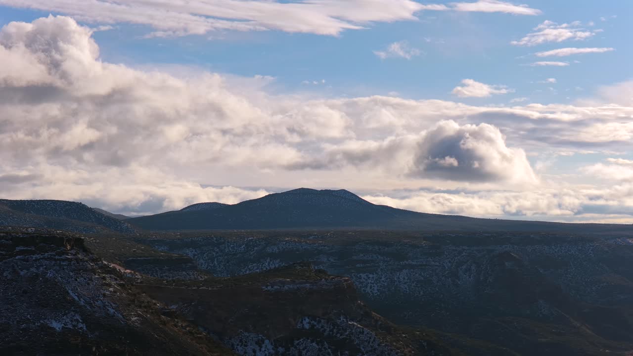 Una ripresa cinematografica laterale cattura un'importante cima innevata che si erge sopra un bordo di altopiano scosceso. Nuvole spettacolari e ampie vedute desertiche definiscono questa aspra natura selvaggia del Sud-ovest.