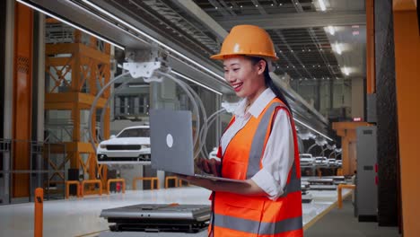 vista lateral de una ingeniera asiática con casco de seguridad trabajando en una computadora portátil mientras está de pie en una fábrica de automóviles ev