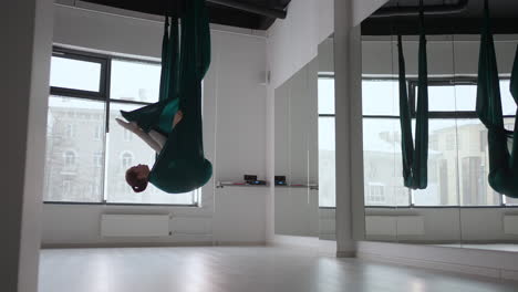 joven mujer hermosa practicando en un columpio de estiramiento aéreo. mujer joven gira su cabeza boca abajo en una hamaca para el yoga aéreo. mujer bastante madura enfocada haciendo yoga aéreo en el gimnasio
