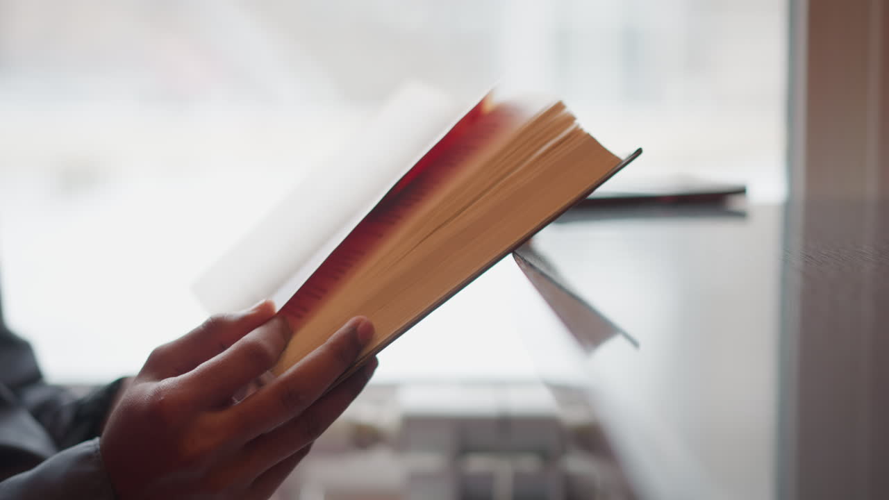 Close-up of hands holding open book in front of window during snowy day, pages turning mid-motion, soft natural light, calm indoor scene with student reading quietly, peaceful focus on printed text