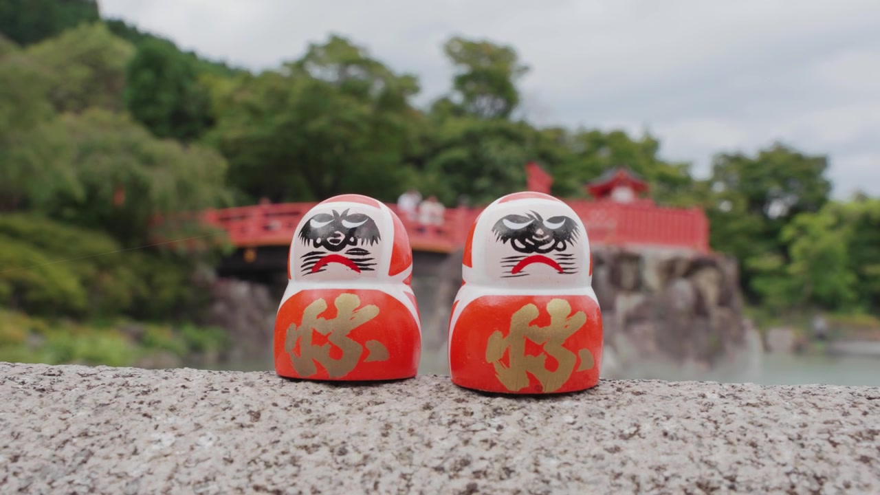 2 røde Daruma-dukker hviler på en stenplatform ved Katsuoji-templet i Osaka, omgivet af frodig grøn vegetation og blød tåge, med en traditionel rød tempelstruktur synlig i baggrunden.