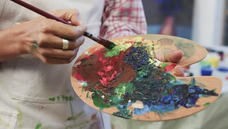 Hands-of-biracial-female-artist-in-apron-mixing-paints-on-palette-using-brush-in-studio,-slow-motion