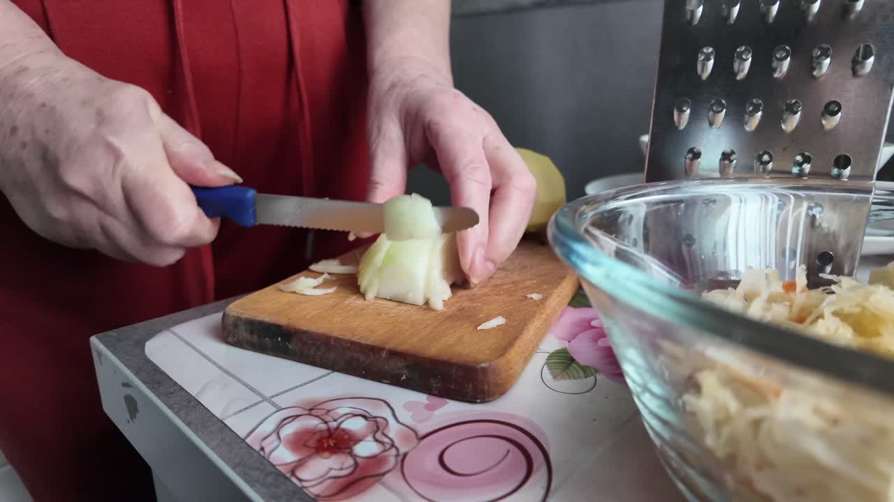 An elderly housewife is cutting an onion into cubes in the kitchen