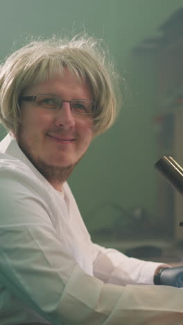 a close-up view of a smiling technician wearing a blonde wig and glasses, working at a laboratory desk with a microscope