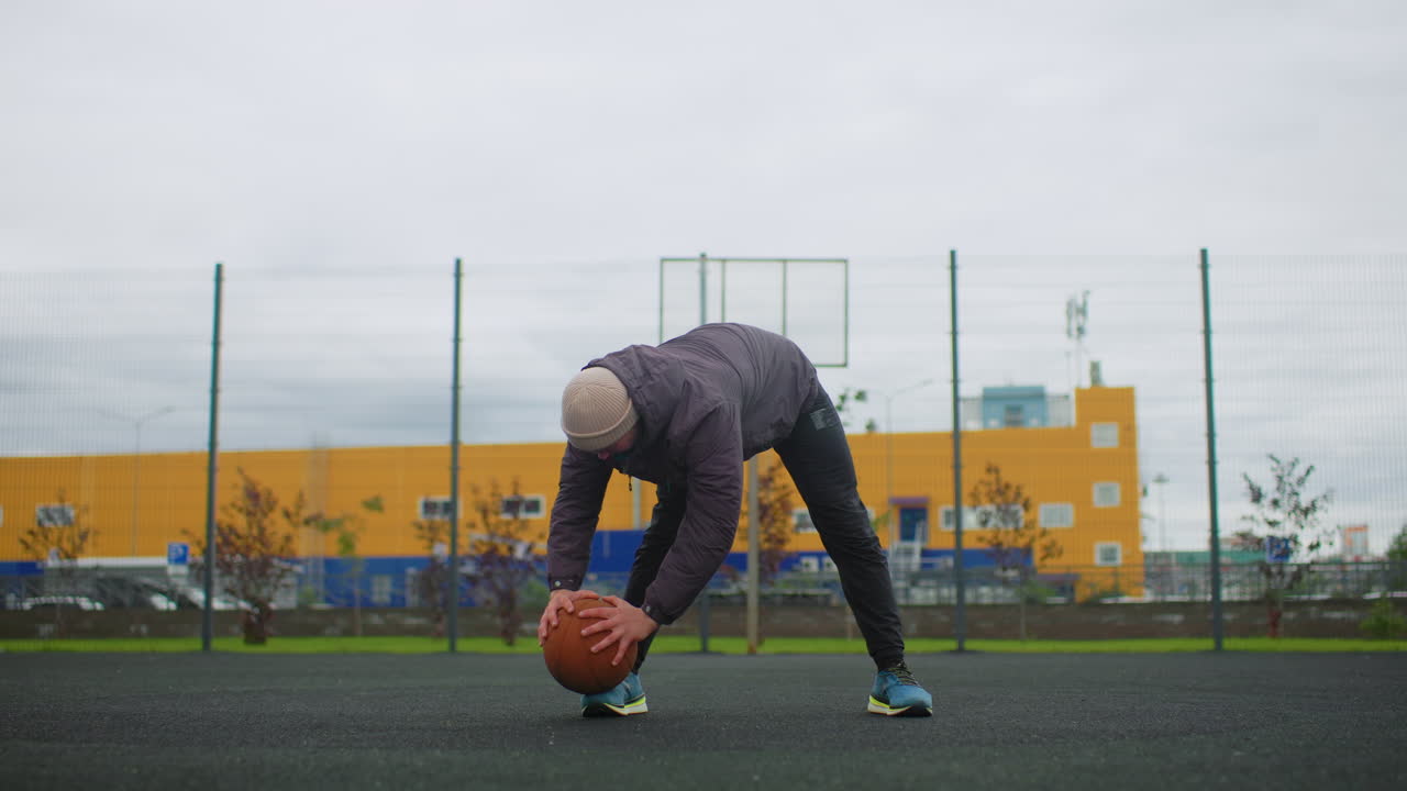 Hvid mand på udendørsbane holder basketball og forbereder opvarmningsrutine under en overskyet himmel ved en indhegnet legeplads i byen. Han er iført jakke og hue, står og bøjer sig for at placere bolden, med en fokuseret holdning