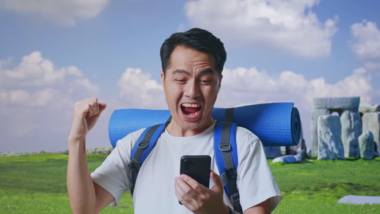 Close Up Of Asian Male Hiker With Mountaineering Backpack Looking At The Smartphone And Screaming Goal Celebrating The Success While Traveling In Stonehenge