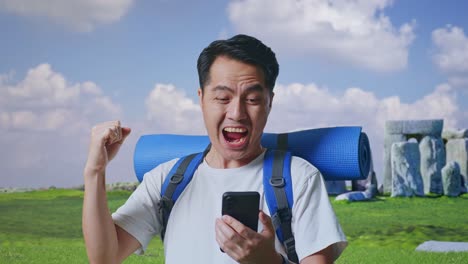 close up of asian male hiker with mountaineering backpack looking at the smartphone and screaming goal celebrating the success while traveling in stonehenge