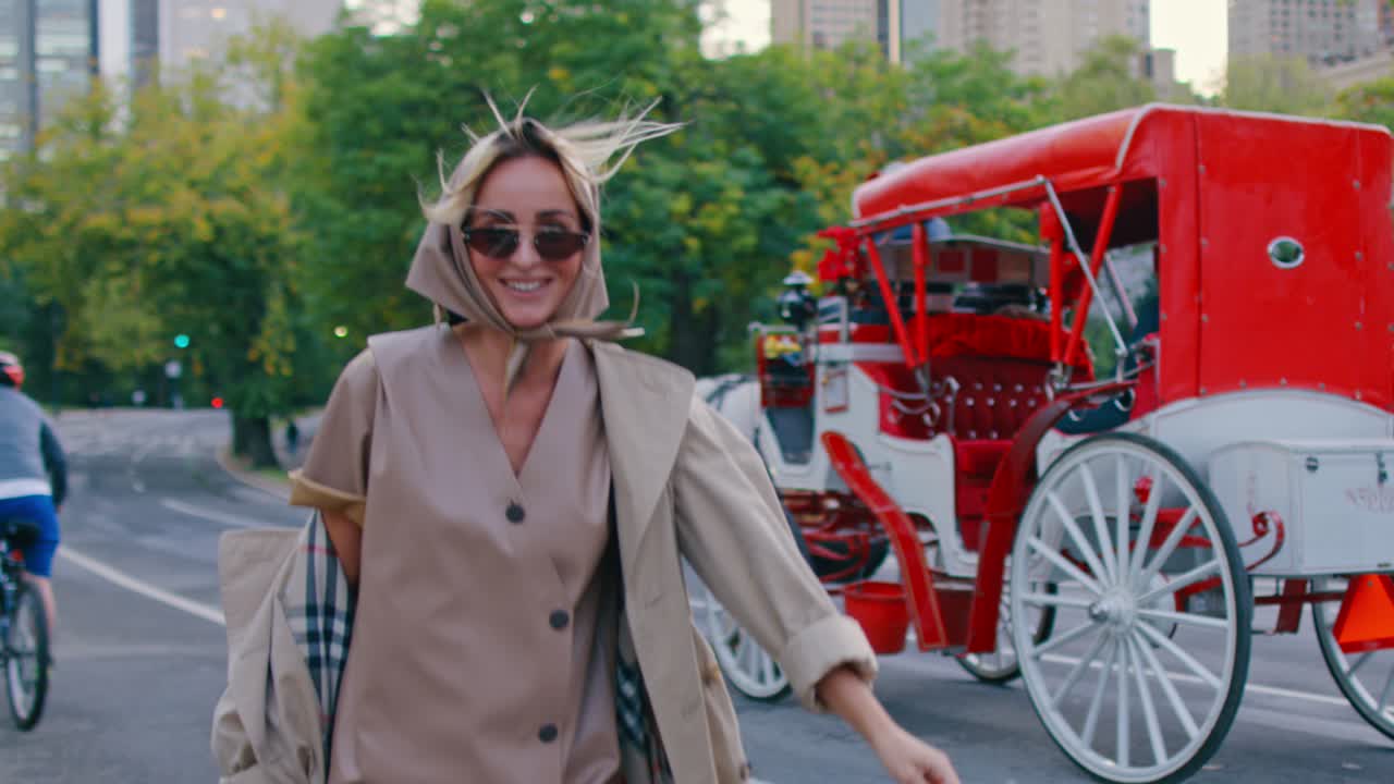 New York, USA, 9 ottobre 2025: Donna sorridente in trench che cammina a Central Park. Donna con foulard e cappotto beige che cammina vicino a una carrozza trainata da cavalli e ciclisti nel parco di New York.