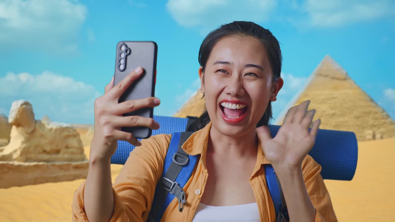 Close Up Of Asian Female Hiker With Mountaineering Backpack Waving Her Hand And Pointing Back While Having A Video Call On Smartphone While Traveling In Pyramid Of Giza