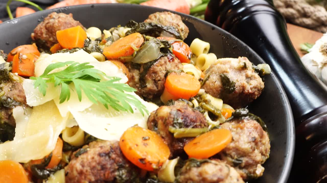 Italian Wedding Soup in a black bowl on a wooden cutting board, close up and panning across the meal. Surrounded by carrots, garlic, onions, vintage tea kettle and a pepper mill. Soup for dinner.