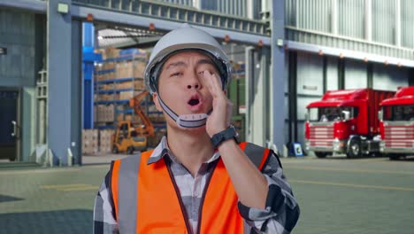 close up of asian male engineer with safety helmet yelling with hand over mouth while standing , outside of logistics distributions warehouse
