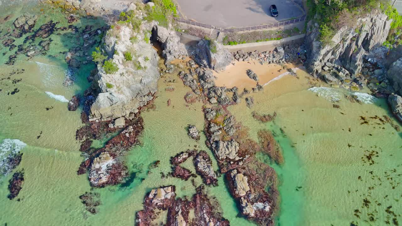 An aerial shot slowly ascending over the stunningly clear turquoise waters and rocky formations of the Uradome Coast in Tottori, Japan, revealing a small sandy beach nestled by the cliffs