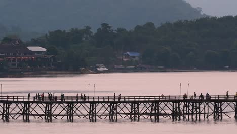 Mon-Bridge-and-a-longboat-followed-speeding-behind-the-structure-while-people-are-walking-on-the-bridge,-silhouetting-as-it-was-getting-dark,-in-slow-motion