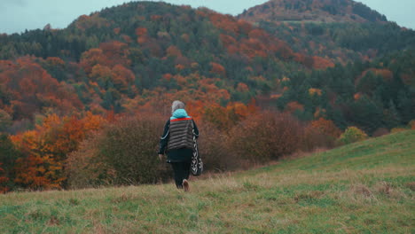 Medium-shot-of-an-older-female-Photographer-with-grey-hair-walking-and-taking-pictures-with-her-camera-in-nature-surrounded-by-orange-coloured-trees-during-a-cold-windy-autumn-day-in-slow-motion