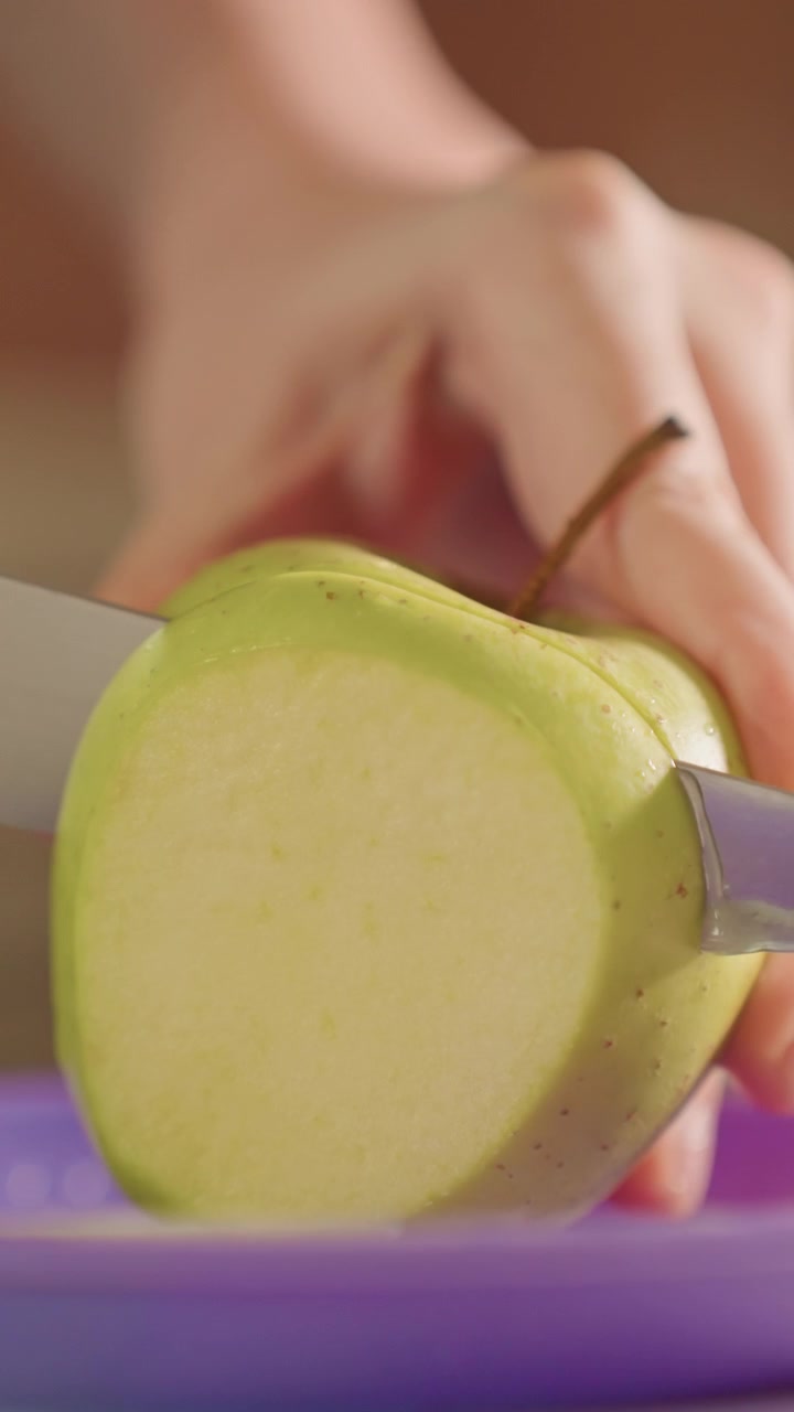 Primo piano di mani che affettano una mela verde a pezzi sottili. Il coltello si muove costantemente attraverso il frutto creando fette lisce. L'ambiente della cucina è implicito con un focus sulla preparazione del cibo.