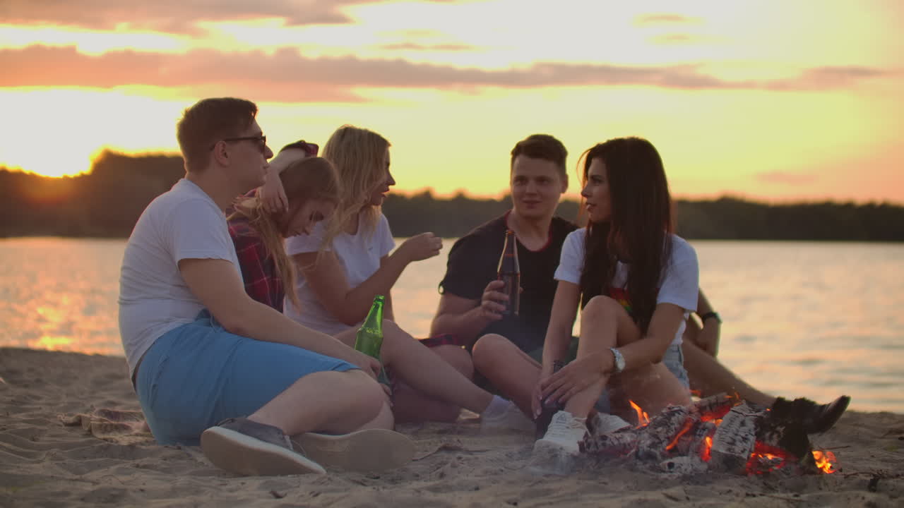 The group of students is sitting in around bonfire on the lake coast. They are spending time cheerfully and they are talking to each other and drinking beer at sunset.