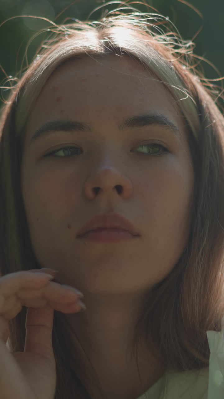 primer plano de una mujer joven con un impermeable translúcido bajando pensativamente su capucha y descansando con los ojos cerrados mientras la luz del sol se filtra suavemente a través de la vegetación circundante