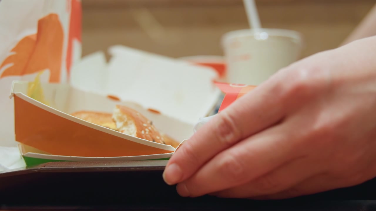 Primo piano di mani che prendono un hamburger su un vassoio, impiegato che recupera un hamburger da asporto dalla confezione di cartone in un ristorante fast casual, panino al sesamo con formaggio fuso e salsa, illuminazione calda del tavolo, dita