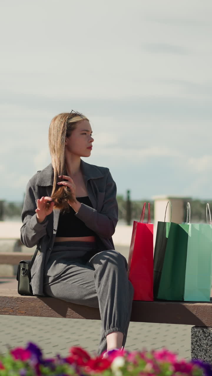 mujer sentada en un banco con tres bolsas de compras de colores a su lado, cruza las piernas mientras se endereza el cabello, mira hacia la distancia en un entorno al aire libre con flores en flor alrededor