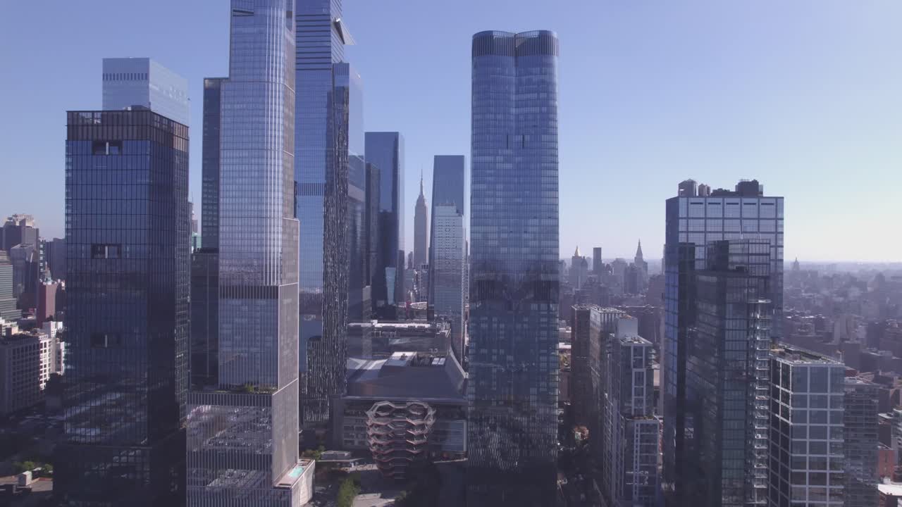 Drone push-in toward The Vessel in Hudson Yards, framed by tall modern skyscrapers under a clear blue sky in Manhattan, New York City